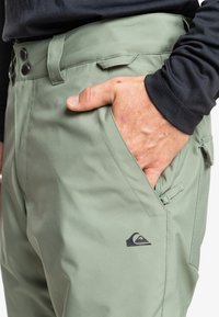 Groene outdoorbroeken met een gladde textuur, dubbele knoopsluiting, zijzak en een klein logo-detail op de bovenbeen.