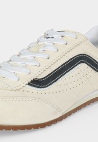 Baskets beiges avec des lacets blancs, une bande noire en forme de vague, de petites perforations et une semelle en caoutchouc marron, vues de côté.