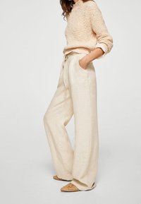 Suéter beige de punto, pantalones de lino beige de corte holgado con bolsillos, y zapatos tipo slip-on de mimbre en tono marrón claro. Textura suave y silueta relajada.