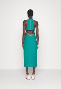 Tyrkysové šaty bez rukávů s odhaleným zadním dílem, zdobené zlatým řetízkem. Šaty jsou těsné, sahají ke kolenům a mají hladký povrch.