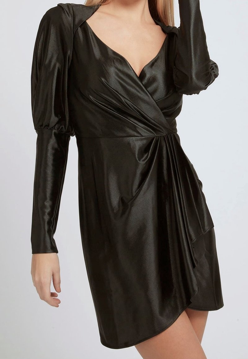 Femme portant une robe portefeuille en satin noir avec de longues manches bouffantes et un décolleté en V profond, debout devant un fond clair uni.