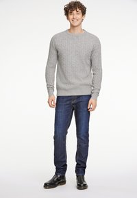 Szary sweter w splot kablowy, niebieskie dżinsy i czarne botki typu chelsea. Modelka stoi na neutralnym tle, uśmiechając się, z krótkimi kręconymi włosami.