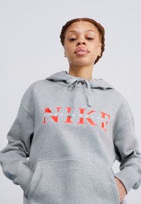 Pilkas džemperis su minkšta medžiaga, priekyje su dideliu oranžiniu ir baltu Nike logotipu, su raišteliais reguliuojama kapuce ir priekinėmis kišenėmis.