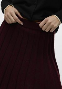 Jupe plissée en tricot bourgogne avec une taille élastique, associée à un cardigan noir avec des boutons ronds et des poignets contrastants.