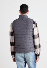 Chaleco gris acolchado con cuello alto, que presenta costuras texturizadas, usado sobre una camisa de cuadros. Jeans de mezclilla visibles y ajuste casual.