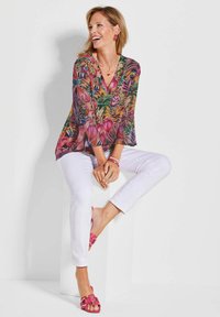 Bloes met bloemenprint en lange, klokvormige mouwen in levendige kleuren. Draagt zij met een witte broek en roze sandalen met een decoratieve gesp.