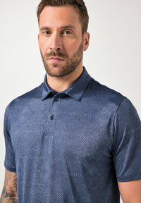 Mann mit kurzen braunen Haaren und Bart, trägt ein blau gemustertes Poloshirt, steht vor einem einfarbigen hellen Hintergrund.