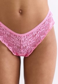Sous-vêtements de style bikini en dentelle rose, avec un motif floral à la taille, présentant un bord festonné et une texture douce visible sur l'ensemble.