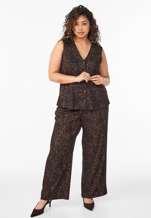 Frau mit lockigem Haar, die ein ärmelloses, dunkel bedrucktes Oberteil mit Leopardenmuster und passende, weit geschnittene Hose sowie schwarze Schuhe trägt, steht vor weißem Hintergrund.