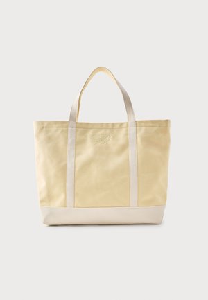 Sac fourre-tout en toile beige avec des anses et un fond crème, présentant un logo brodé subtil au centre à l'avant.