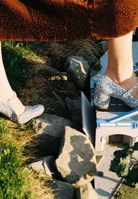 Zapatos de tacón Mary Jane brillantes en plata con tacones gruesos y correas al tobillo, colocados sobre piedras al lado de un pequeño modelo de edificio. Césped al fondo.
