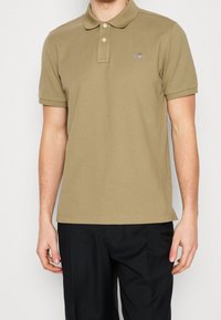 Khaki polo tričko s límečkem, krátkými rukávy a malým logem. Tkanina vypadá hladce, má rovný střih a minimální detaily.