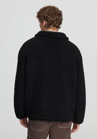 Giacca in pile nera con colletto alto, maniche lunghe e forma squadrata. Texture morbida, dettagli minimi e design casual.