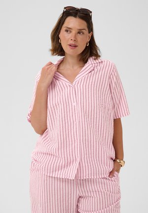 Donna con capelli castani corti indossa una camicia a maniche corte a righe rosa e bianco con bottoni e pantaloni abbinati, occhiali da sole in testa, orecchini e bracciale d'oro.