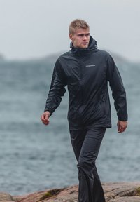 Sort vandtæt jakke med hætte, lynlås foran og elastiske manchetter, parret med matchende sorte vandtætte bukser. Regn-synlig overfladetekstur.