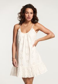 Bijela pamučna haljina s višeslojnim volanima na rubu, koja ima cvjetne izreze u broderie anglaise stilu, tanke naramenice i opušteni kroj.