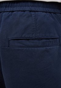 Pantalones azul marino con cinturilla elástica y un bolsillo trasero con detalle en relieve "MUSTANG". La tela tiene un acabado texturizado.