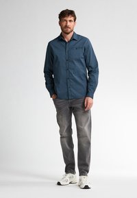 Camicia button-up di colore blu scuro con motivo geometrico, abbinata a jeans di denim grigi e sneakers bianche, in piedi su uno sfondo neutro.