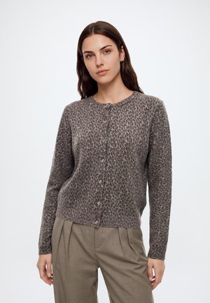 Woman with long brown hair wearing a brown leopard print buttoned cardigan and taupe pleated trousers against a plain background.