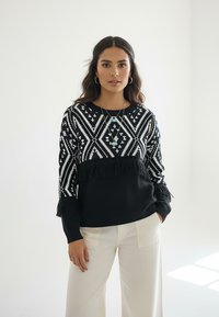 Czarnobiały dzianinowy sweter z wzorem diamentowym, z frędzlami na dole i rękawach, z okrągłym dekoltem.