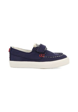 Zapatillas de lona azul marino tipo slip-on con suela de goma blanca, con detalles en rojo en la costura y una correa de cierre con gancho y bucle. Superficie texturizada.