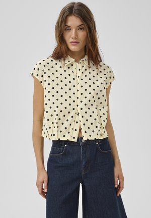 Jonge vrouw draagt een crème kleurig shirt met korte mouwen en zwarte polkadots, gecombineerd met een donkere, high-waisted spijkerbroek, tegen een effen achtergrond.