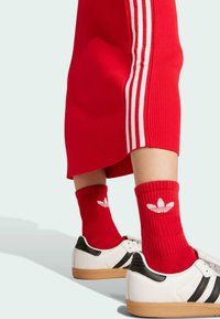 Baskets blanches avec des accents noirs et semelles en gomme, accompagnées de chaussettes à côtes rouges arborant un logo Adidas blanc, et d'une jupe rouge avec des rayures blanches.