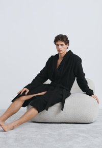 Melna halāts ar kapuci, garām piedurknēm un jostu. Sēž uz mīkstas, gaišas, apaļas krēsla uz pelēka paklāja. Gluda tekstūra.