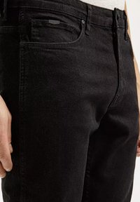 Czarne dżinsy mają slim fit, widoczne szlufki na pasek oraz przednią kieszeń z małą metką. Tekstura jest gładka z delikatnym wyblaknięciem.