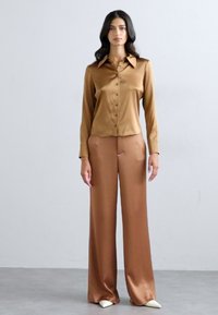 Chemise longue à manches longues en satin marron avec col pointu et boutons, associée à un pantalon large assorti. Texture lisse et coupe ajustée.