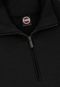 Black zip-up sweater with ribbed collar, featuring a metal zipper and a logo tag. Fabric appears smooth with a soft texture.