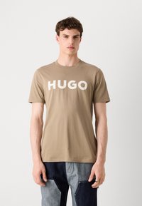 Beige tričko vyrobené z bavlny, s kulatým výstřihem a výrazným bílým nápisem "HUGO" na přední straně. Krátké rukávy, volný střih.
