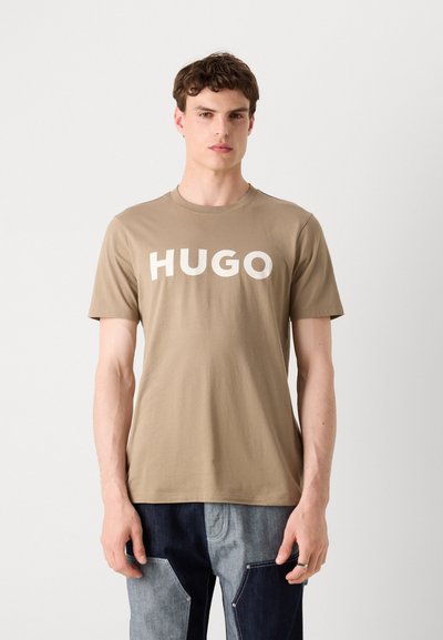 Бежова тениска от памук с кръгло деколте и смело бяло „HUGO“ на предната част. Къси ръкави, свободна кройка.