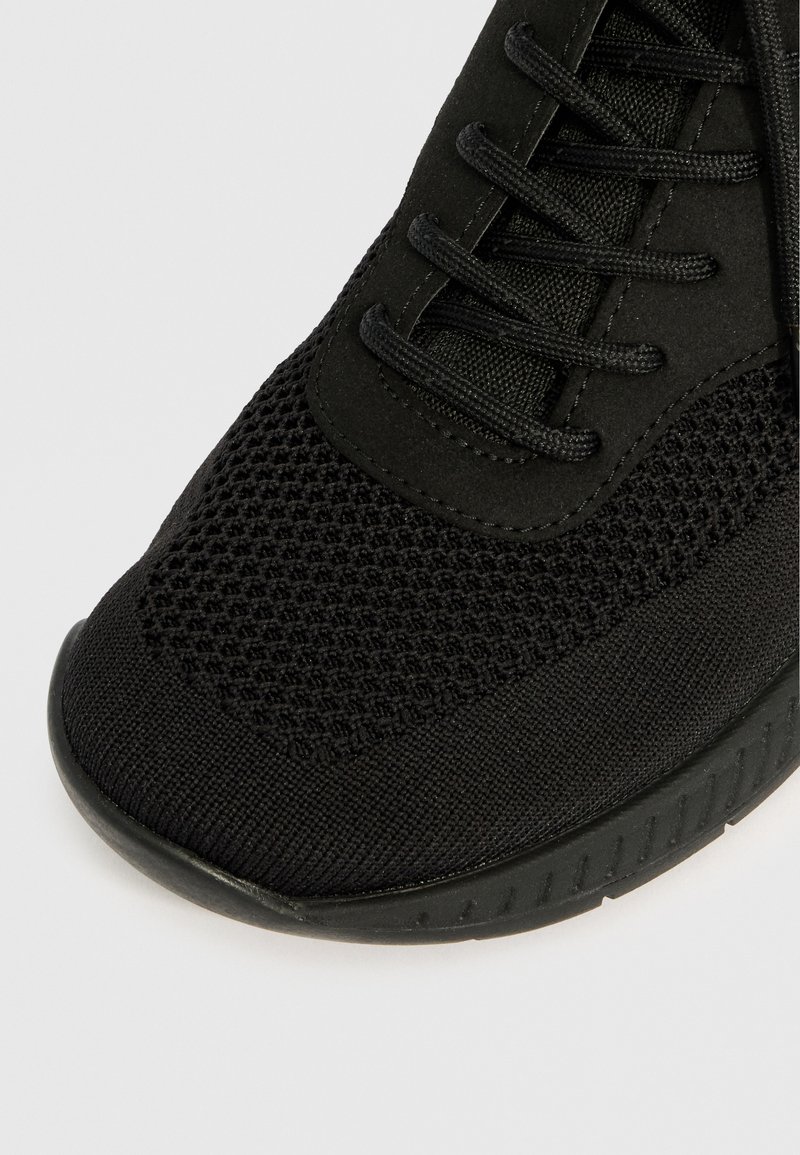 Close-up van de voorkant van een zwarte sneaker met gebreide mesh-stof, gevlochten veters en een gestructureerde rubberen zool op een lichte achtergrond.