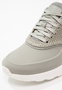 Sneaker grigio chiaro con parte superiore liscia, dettagli in rete, lacci piatti e suola bianca. Il materiale ha una finitura opaca e accenti testurizzati.