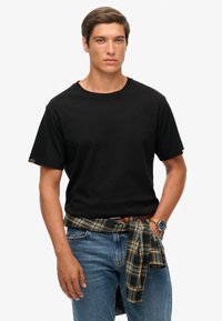 Sort kortærmet t-shirt med rund halsudskæring og diskret logo-detalje. Kombineret med blå jeans og en ternet skjorte bundet om taljen.