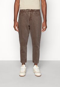 Jean en denim marron avec cinq poches, passants pour ceinture et coupe fuselée. Porté avec des baskets blanches et beiges pour un look décontracté.