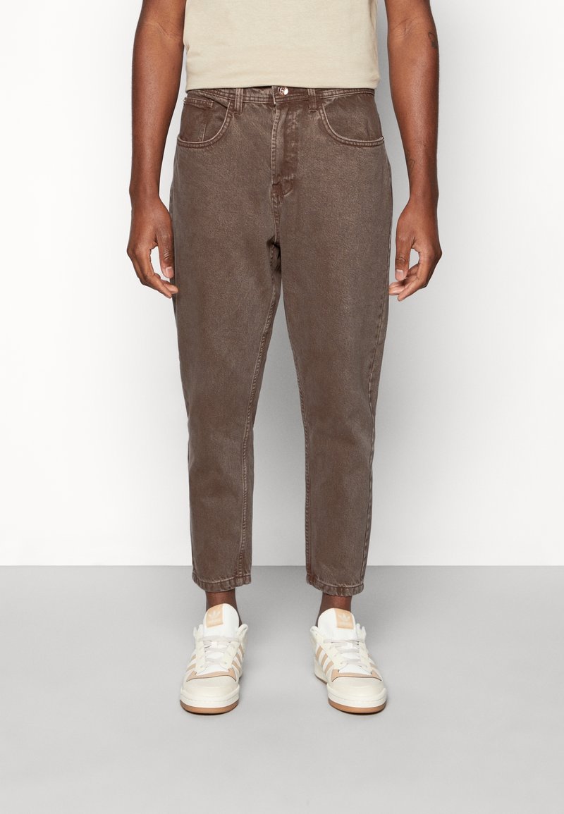 Jean en denim marron avec cinq poches, passants pour ceinture et coupe fuselée. Porté avec des baskets blanches et beiges pour un look décontracté.