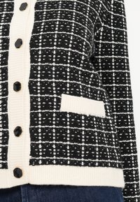 Κουβέρτα σε μαυρό άσπρο σχέδιο με ριχτό υλικό, που διαθέτει κρεμ μπροστινό μέρος, μεγάλους μαύρους κουμπιά και μια μικρή τσέπη στην αριστερή πλευρά.