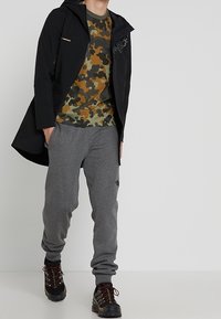 Giacca impermeabile nera con cappuccio, camicia a motivo mimetico in verde e marrone, pantaloni della tuta grigi e stivali da escursionismo scuri con accenti metallici.