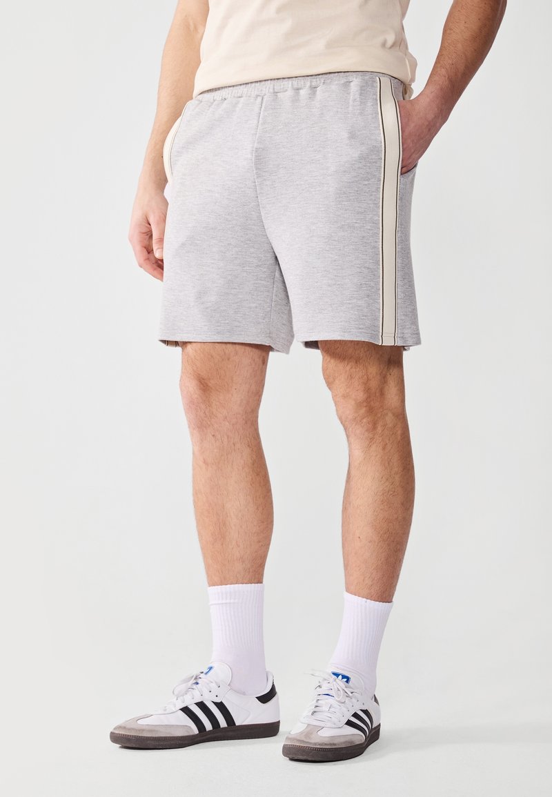 Shorts en coton gris avec une taille élastique, des poches latérales et des bandes latérales crème. Portés avec des baskets blanches et noires et des chaussettes blanches.
