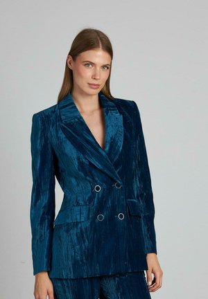 Blazer de doble botonadura en terciopelo verde profundo, con solapas en pico, hombros estructurados y botones en tono plateado. Textura suave con brillo.