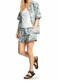 Dwuczęściowy zestaw składający się z koszuli z krótkim rękawem w palmowy wzór oraz dopasowanych szortów w jasnych kolorach. Stylizację dopełniają sandały z czarnymi paskami.