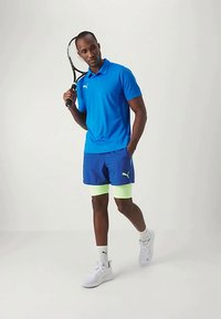 Urheiluasu, joka koostuu sinisestä polo-paidasta sekä sinisistä shortseista vihreillä yksityiskohdilla. Täydennettynä valkoisilla tennarit ja kädessä tennismaila.