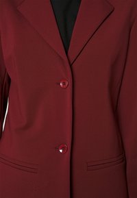 Blazer rouge foncé avec deux boutons assortis, revers crantés et deux poches fendues horizontales, porté sur un haut noir.