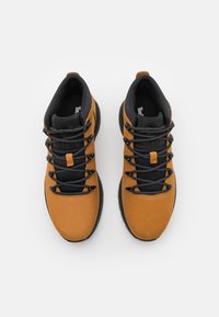 Bocanci de drumeție din suede maro, cu șireturi negre și limbă din plasă, având o talpă din cauciuc negru și branding Timberland pe lateral.