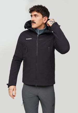 Man with dark curly hair and mustache adjusts the hood of a black Mammut jacket, wearing grey pants, standing against a plain light background.