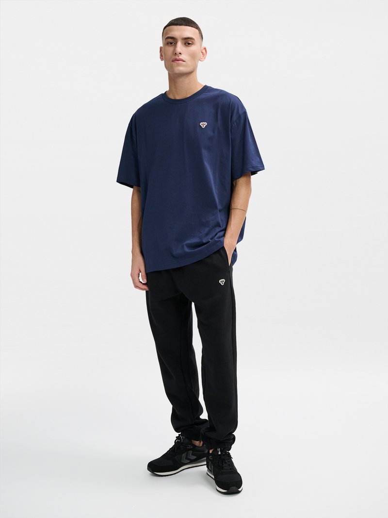 Ung mand iført navy oversized t-shirt og sorte joggers med logoer, iført sorte sneakers, foran en ensfarvet hvid baggrund.