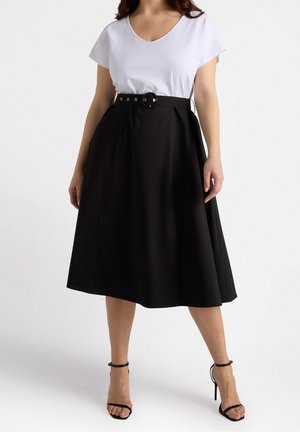 Woman wearing a white short-sleeve V-neck top tucked into a high-waisted black A-line skirt with a black belt and black ankle-strap heels.