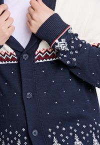 Marinblå cardigan med röda och vita mönstrade axelaccenter, med knappar och en texturerad stickning. Dekorerad med snöflinge- och trämotiv.